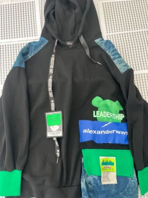 Svart hoodie Alexander Wang x Undercover - Unik svart hoodie med gröna och blå detaljer från Alexander Wang x Undercover. Stora tryck med texten 'LEADERSHIP' och 'alexanderwang', samt patchar och denimliknande partier på axlar och ficka. Huva med snörning och ribbade muddar i grönt. Perfekt för dig som gillar streetwear.