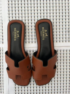 Bruna Härmes Åran sandaler - Säljer ett par klassiska bruna Härrmès Åran sandaler i läder med den ikoniska H-designen. Sandalerna har svart innersula och låg klack. Perfekta för dig som gillar stilrena och lyxiga detaljer från ett exklusivt märke.