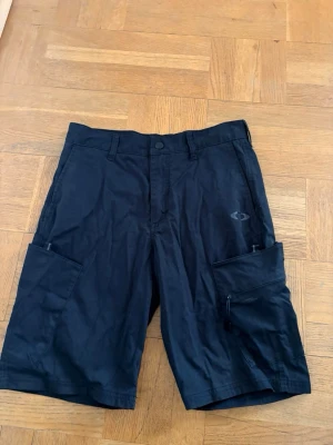 Svarta cargoshorts från Oakley - Säljer ett par svarta cargoshorts från Oakley med flera praktiska fickor, inklusive en med dragkedja. Shortsen har normal passform, bälteshällor och knappstängning. Perfekta för dig som gillar funktionella och stilrena plagg. Perfekta friluftsshort eller slit å släng shorts. Nypris 700kr mitt pris 350kr. Storlek 30 vilket motsvarar S/M