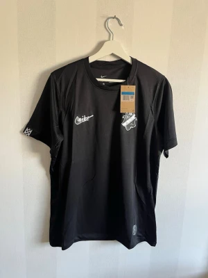 Svart AIK x Nike t-shirt - Snygg svart t-shirt från Nike i samarbete med AIK och Smoking Culture. Tröjan har AIK-logga och Nike-logga på bröstet, samt schackrutiga detaljer på ärmarna. Tillverkad i ett lätt och funktionellt material, perfekt för träning eller sport.