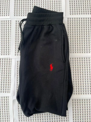 Svarta mjukisbyxor från Polo Ralph Lauren - Svarta mjukisbyxor från Polo Ralph Lauren med röd broderad logga på benet. Byxorna har resår i midjan med dragsko och sidofickor. Tillverkade i mjukt bomullsmaterial för maximal komfort och avslappnad stil.