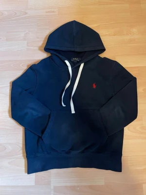 Marinblå hoodie från Polo Ralph Lauren - Klassisk marinblå hoodie från Polo Ralph Lauren med vit dragsko och röd broderad logga på bröstet. Hoodien har känguruficka framtill och ribbade muddar vid ärmar och nederkant. Perfekt för en avslappnad och stilren look. Har ett hål annars i mycket bra skick! Priset kan diskuteras! Först till kvar!!