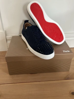 Christian Loubotin skor - Säljer ett par marinblå sneakers från Christian Louboutin med coola svarta nitar på tån, klassisk röd sula och vita kanter. Skorna har snörning och är tillverkade i mocka för en lyxig känsla. Perfekta för dig som vill sticka ut med din stil.