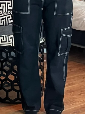 Svarta cargobyxor med kontrastsömmar - Säljer ett par svarta cargobyxor med vita kontrastsömmar och flera stora fickor på sidorna. Byxorna har rak passform och en cool streetwear-vibe.