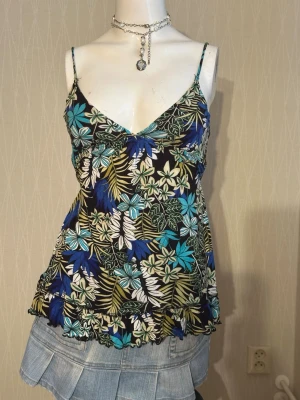 Blommig linnetopp med volangkant - Snygg linnetopp med tunna axelband och blommigt mönster i blått, grönt, vitt och gult på svart botten. Toppen har v-ringning och volangkant nedtill, perfekt för sommarens alla outfits.