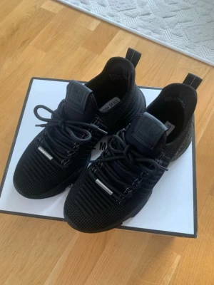 Svarta sneakers från Steve Madden - Snygga helsvarta sneakers från Steve Madden med stickad ovandel och tjock sula. Skorna har svarta skosnören med metalländar och en sportig, modern look. Perfekta för dig som gillar streetstyle och vill ha bekväma skor med cool design. Använda en gång. Mindre i storlek. 