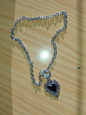 Silverfärgat halsband med svart hjärthänge - Unikt halsband i silverfärg med en grov kedja och ett detaljerat hänge i form av ett svart hjärta omgivet av dekorativa mönster. Hänget har en vintageinspirerad look med små pärlliknande detaljer runt hjärtat. Perfekt för dig som gillar statement-smycken.