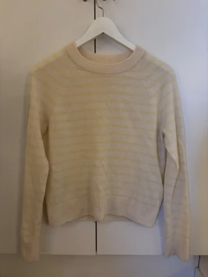 Cashmere tröja Samsoe & Samsoe - Mysig stickad cashmere tröja i ljusbeige och vitt med diskreta ränder. Tröjan har rund halsringning, långa ärmar och ribbade muddar vid ärmslut och nederkant. Ett litet hål vid ena ärmen. 