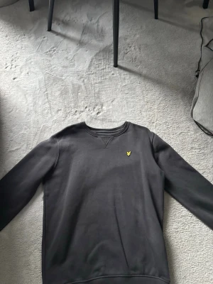 Svart sweatshirt från Lyle & Scott - Klassisk mörkgrå sweatshirt från Lyle & Scott Junior med rund halsringning och gul broderad logga på bröstet. Tröjan har ribbade muddar vid ärmslut och nederkant. Perfekt för en avslappnad och stilren look.