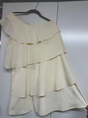 Offshoulder vit omlottklänning - Snygg offshoulder klänning i vit ton med flera lager och volanger. Klänningen har en omlott-design och är perfekt för dig som vill ha en unik och trendig look. Materialet känns lätt och passar bra till varma dagar.