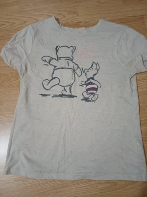 Grå T-shirt med Nalle Puh och Nasse - Säljer en ljusgrå T-shirt med tryck av Nalle Puh och Nasse på ryggen. Motivet är tecknat i svart med rosa detaljer på Nasses tröja. T-shirten har korta ärmar och är i mjukt bomullsmaterial. Perfekt för dig som gillar klassiska Disney-karaktärer.