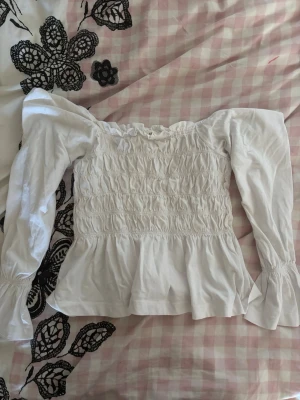 Vit offshoulder blus med volang - Trendig vit offshoulder blus med långa puffärmar och smockad överdel. Blusen har volang nedtill och resår vid ärmslut för en snygg siluett. Perfekt för dig som gillar romantisk och feminin stil.