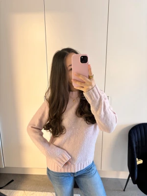 Ljusrosa stickad tröja - Mysig ljusrosa stickad tröja med rund hals och långa ärmar. Tröjan har ribbade muddar vid ärmslut och nederkant, vilket ger en skön och avslappnad passform. Perfekt att matcha med jeans för en enkel och stilren look.