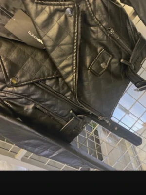 Svart quiltad skinnjacka Fashion Nova - Snygg svart skinnjacka från Fashion Nova med quiltat mönster, dragkedjor, bred krage och bälte i midjan. Jackan har flera fickor med dragkedja och coola detaljer som ger en edgy look. Perfekt för dig som vill sticka ut med din stil.