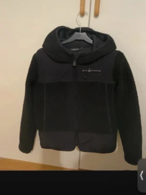 Svart teddyjacka från Sail Racing - Mysig svart teddyjacka från Sail Racing med huva och hel dragkedja. Jackan har en bröstficka med dragkedja och logotyp på vänster sida. Materialet är fluffigt och mjukt, perfekt för kalla dagar. Snygg och enkel design som är lätt att matcha.