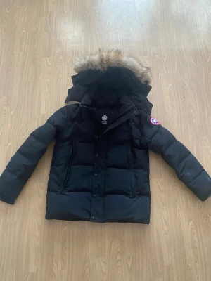 Svart dunjacka från Canada Goose - Säljer en svart dunjacka från Canada Goose med avtagbar huva och pälsdetalj. Jackan har en klassisk patch på ärmen, dragkedja och knappar framtill samt två stora fickor. Perfekt för kalla vinterdagar och riktigt varm.