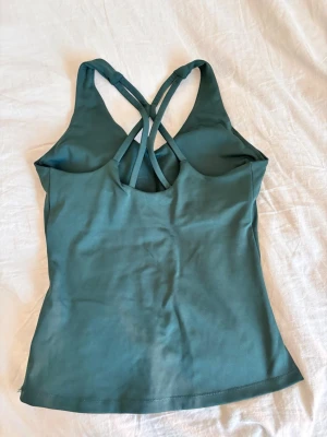 Mörkgrön linnetopp med korsade band - Snygg mörkgrön linnetopp med smala axelband som korsas i ryggen. Toppen är ärmlös och har en v-ringad halsringning. Perfekt för träning eller sommarens varma dagar. Tillverkad i mjukt syntetmaterial för skön passform.