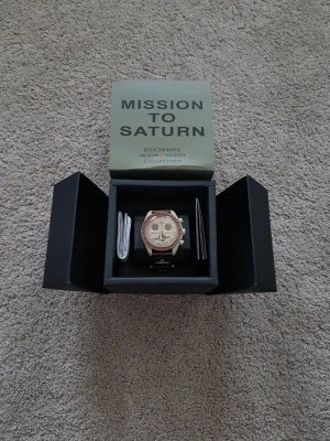 Omega x Swatch Mission to Saturn - Säljer en Omega x Swatch Speedmaster MoonSwatch Mission to Saturn. Klockan har en beige och brun boett i biokeramik, brun bezel och matchande brunt band med Omega-logga. Snygg kronografdesign med Saturnus-detalj på urtavlan. Kommer i originalbox.