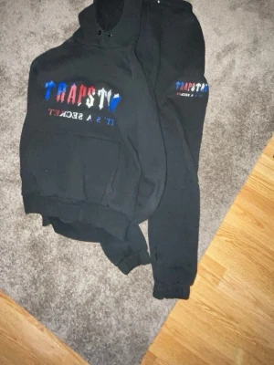 Svart TRAPSTAR loungewear set - Säljer ett svart loungewear set från Trapstar med färgglad logga i blått, rött och vitt på både hoodie och byxor. Hoodien har magficka och byxorna har resår i midjan och vid bensluten. Perfekt för chill dagar hemma.