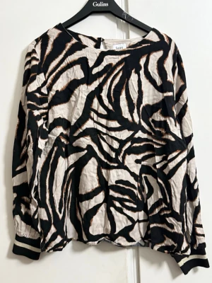Zebramönstrad blus från Saint - Snygg blus från Saint med zebramönster i svart, beige och bruna detaljer. Blusen har rund halsringning, långa ärmar med svarta muddar och knäppning bak i nacken. Tillverkad i ett lätt, skrynkligt tyg som ger en avslappnad look.