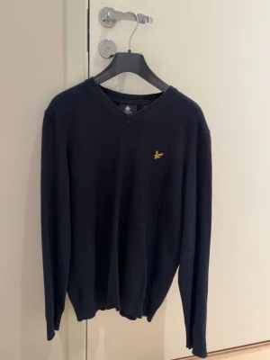 Mörkblå stickad tröja Lyle & Scott - Snygg mörkblå stickad tröja från Lyle & Scott med V-ringning och den klassiska gula logon broderad på bröstet. Tröjan har långa ärmar och ribbade muddar vid ärmslut och nederkant. Perfekt till lager på lager och enkel att matcha.