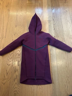 Lång mörklila jacka med huva - Lång mörklila jacka med hel dragkedja framtill och huva. Jackan har en sportig look med svarta detaljer längs bröstet och är tillverkad i ett mjukt syntetmaterial. Perfekt för kyliga dagar och har lång ärm för extra värme.