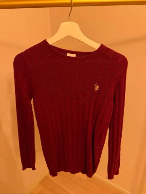 Vinröd stickad tröja U.S. Polo Assn. - Vinröd stickad tröja från U.S. Polo Assn. med diskret logga broderad på bröstet. Tröjan har rund halsringning, smal passform och ribbade muddar. Perfekt för lager på lager och ger en klassisk look.