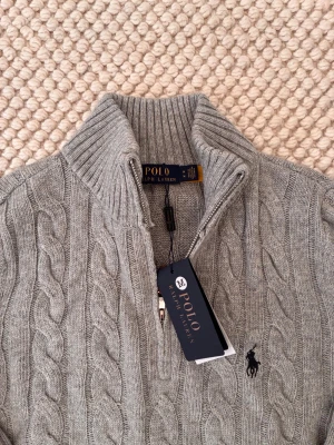 Grå kabelstickad tröja Polo Ralph Lauren - Snygg grå kabelstickad tröja från Polo Ralph Lauren med halv dragkedja och hög krage. Tröjan har ribbade muddar och nederkant samt den klassiska broderade loggan på bröstet. Perfekt för dig som gillar stilren och tidlös design.