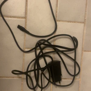 Svart laddare till babyliss - En svart laddare med lång sladd för mobiltelefon. Laddaren har en klassisk stickkontakt och en rundad kontakt i andra änden, vilket passar äldre mobilmodeller. Tillverkad i plast med enkel design och smidig att ta med sig.