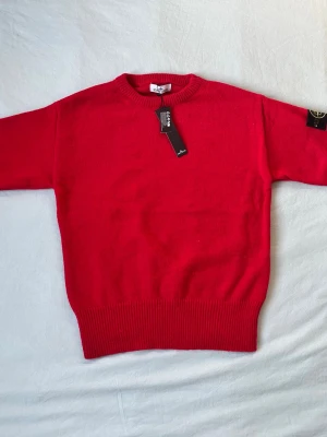 Röd stickad tröja från Stone Island - Snygg röd stickad tröja från Stone Island med klassisk rund halsringning och ribbade muddar. Tröjan har den ikoniska Stone Island-loggan på ärmen och är tillverkad i mjukt material som passar perfekt till höst och vinter.