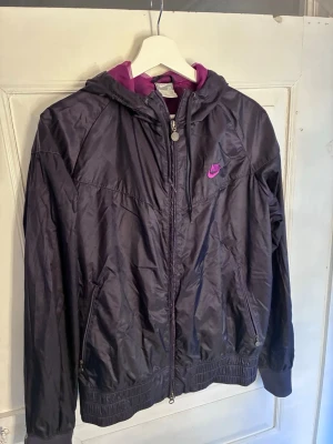 Mörklila vindjacka från Nike - Nike vindjacka i mörklila meshfoder och lila Nike-logga på bröstet. Jackan har huva, dragkedja framtill, två fickor med dragkedja och ribbade muddar. Materialet är glansigt och lätt, perfekt för blåsiga dagar. Passar snarare M/L