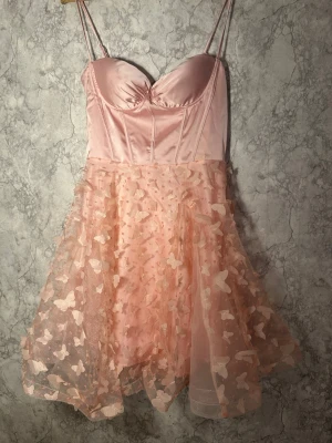 Rosa kort klänning med fjärilar - Supersöt rosa kort klänning med smala axelband och byst i glansigt satinliknande material. Kjolen är i flera lager med 3D-applikationer av fjärilar och ger en fluffig look. Baksidan har snörning för extra detalj och passform. Perfekt för dig som vill sticka ut!