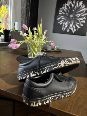 Svarta sneakers från Zara med graffiti-detaljer - Unika svarta sneakers från Zara med cool graffiti-inspirerad text i vitt runt sulan. Skorna har en klassisk låg profil, svarta skosnören och är tillverkade i slätt konstläder. Perfekta för dig som vill sticka ut med en edgy streetstyle.