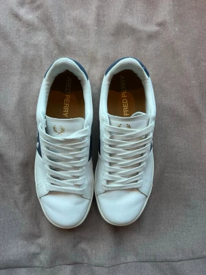 Vita Fred Perry sneakers med blå detalj - Snygga vita sneakers från Fred Perry med marinblå sidopanel och broderad lagerkrans. Skorna har klassisk låg profil, vita snören och detaljer i läder. Perfekta för en clean och stilren look.