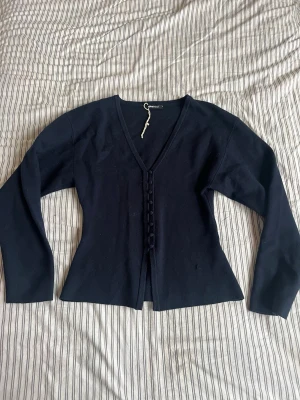 Mörkblå kofta från Gina Tricot - Säljer en mörkblå stickad kofta från Gina Tricot med v-ringning och knappar framtill. Koftan har långa ärmar och en smickrande, något figurnära passform. Perfekt att slänga över en topp eller klänning för extra stilpoäng.