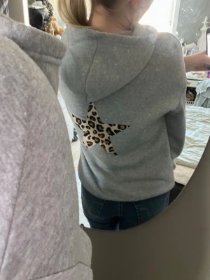 Grå hoodie med leopardstjärna - Snygg grå hoddie. På ryggen finns en stjärna i leopardmönster som ger en cool kontrast till den enkla designen. Perfekt för dig som gillar streetstyle och vill sticka ut lite extra. Mjuk och skön, passar till jeans eller leggings.