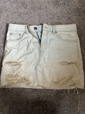 Ljusblå jeanskjol med slitningar - Cool ljusblå jeanskjol med slitna detaljer och rå kant längst ner. Kort, i strl 36/S