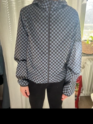 Gråblå rutmönstrad vindjacka - Snygg vindjacka med gråblått rutmönster och huva. Jackan har dragkedja framtill och är tillverkad i ett lätt material som passar perfekt för blåsiga dagar. 