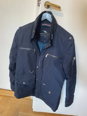 Mörkblå jacka från Snoot - Snygg mörkblå jacka från Snoot med flera fickor, dragkedjor och knappar. Jackan har hög krage och coola detaljer som metallringar och bröstfickor. Perfekt för dig som gillar stilren och funktionell design.