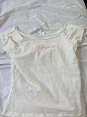 Vit t-shirt Ma chérie Gina Tricot - Söt vit t-shirt från Gina Tricot med rosa texten 'Ma chérie' på bröstet. T-shirten har korta ärmar och en klassisk passform. Tillverkad i mjuk bomull, perfekt för en avslappnad och trendig look.