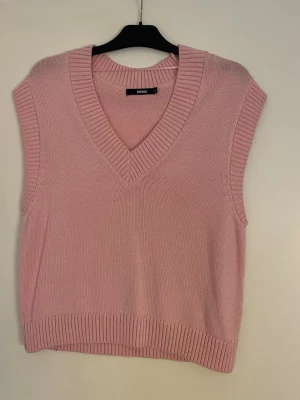 Ljusrosa stickad väst från BikBok - Säljer en ljusrosa stickad väst från BikBok med bred ribbad v-ringning och ribbade kanter. Västen är ärmlös och har en avslappnad passform, perfekt att styla över en skjorta eller t-shirt för en trendig look.