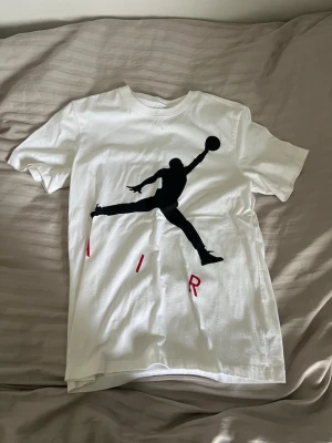 Vit Air Jordan t-shirt - Säljer en vit t-shirt från Jordan med den ikoniska Jumpman-loggan i svart på framsidan och texten AIR i rött. Klassisk passform och rund halsringning. Perfekt för dig som gillar streetwear och sportig stil.