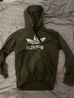 Grön Adidas hoodie med logga - Mörkgrön hoodie från Adidas med stor vit Trefoil-logga och text på bröstet. Klassisk känguruficka framtill och justerbar huva med svarta snören. Tillverkad i mjukt bomullsmaterial, perfekt för en avslappnad streetwear-look.