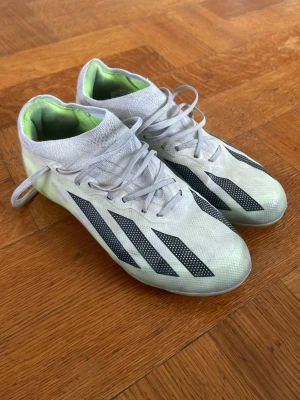 Vita Adidas fotbollsskor med svarta ränder - Säljer ett par vita Adidas fotbollsskor med klassiska svarta ränder på sidorna och limegröna detaljer på insidan. Skorna har snörning och en strumpliknande krage för extra stöd. Tillverkade i syntetmaterial som ger bra grepp och känsla på planen. Skorna har blivit lite upprivna på ena sulan men märks inte när man spelar.