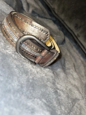 Bälte med bling - Snyggt brunt bälte i skinn med silverfärgade nitar och glittriga detaljer längs hela bandet. Bältet har en robust metallspänne och passar perfekt för att ge outfiten lite extra edge. 