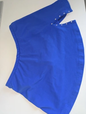 Blå kortkjol med strasstenar - Snygg blå kortkjol i mjukt bomullsmaterial med strasstenar vid linningen och en liten dekorativ spiral i strass baktill. Kjolen har en enkel, utsvängd form och italiensk flaggdetalj vid midjan. Perfekt för en färgstark och lekfull stil. Konståkning kjol