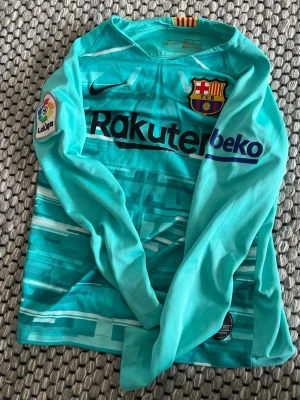 Turkos FC Barcelona tröja Nike M - Säljer en turkos långärmad FC Barcelona matchtröja från Nike med Dri-Fit material. Tröjan har klubbmärke, La Liga-märke på ärmen och sponsorloggor på bröstet. Snygg design med grafiskt mönster och gula/röda detaljer i nacken.
