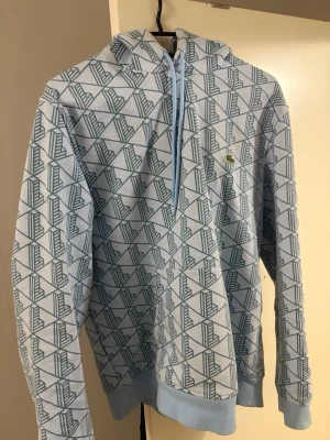 Mönstrad hoodie från Lacoste - Snygg hoodie från Lacoste i ljusblått med geometriskt mönster över hela plagget. Klassisk huva med snörning och ribbade muddar. Liten Lacoste-logga på bröstet. Perfekt för dig som gillar streetwear och vill sticka ut lite extra. Nästan helt ny säljer för den är liten på mig ord pris 1800
