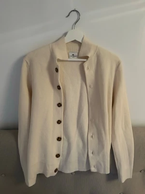 Beige kofta från John Henric - Stilren beige kofta från John Henric med långa ärmar och bruna knappar framtill. Koftan har en rak passform och ribbade muddar vid ärmslut och nederkant. Perfekt för lager-på-lager och enkel att matcha med olika outfits. Kontakta vid eventuella frågor😁