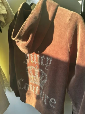 Brun Juicy Couture hoodie med strass - Snygg brun hoodie från Juicy Couture med huva och glittrande strassdetaljer på ryggen. Texten 'Juicy Couture' och en krona i strass ger en lyxig och trendig vibe. Perfekt för dig som gillar statement-plagg och vill sticka ut.